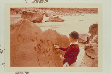 Dock Marston marks for the photographers the inscription made by Harry McDonald of the Best Expedition after they locked a boat on the flat rock at upper left on July 21, 1891. Rapid No. 16 at Mile 204 1/2